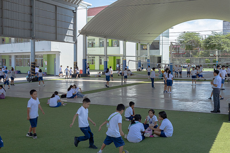 Dozens of children in school uniforms play and socialize under a large covered area in a school courtyard with artificial grass and concrete flooring.