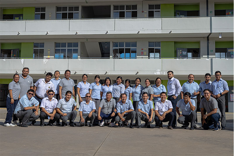 A group of school staff members pose for a photo in front of a multi-story school building with balconies and classrooms.