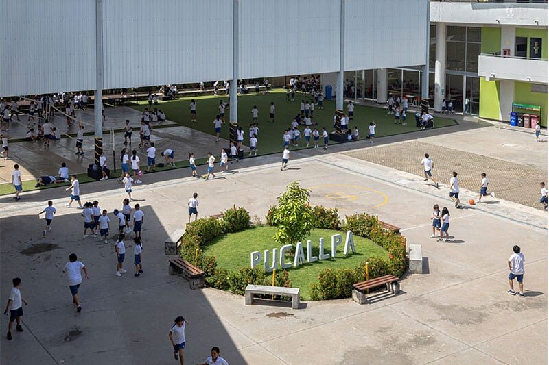 Children in school uniforms play and socialize in a large, open school courtyard with a green area marked “Pucallpa” in the center.