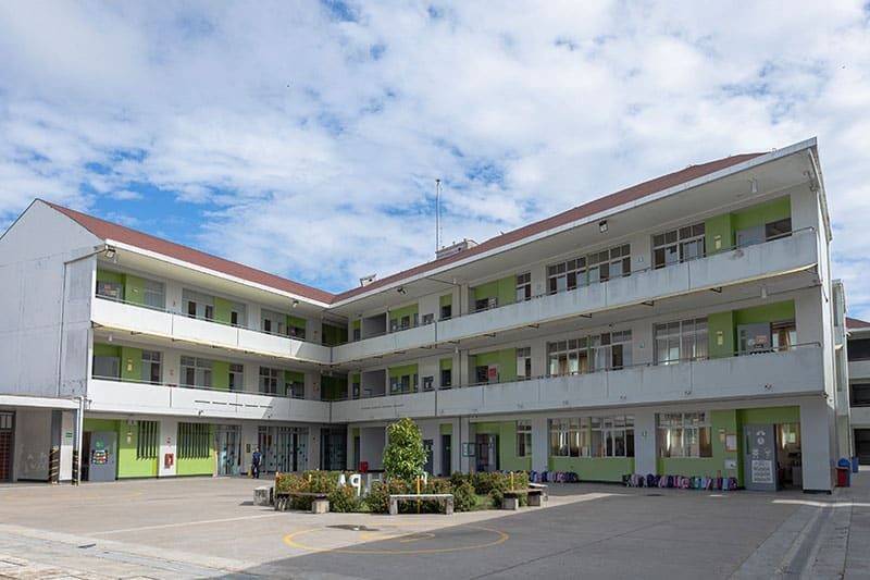 A modern three-story school building with green accents surrounds an open courtyard with a central landscaped area and student backpacks lined up along the ground floor.