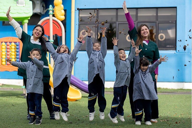 Two teachers and a group of young children joyfully playing and throwing leaves in the air on a playground outside a school building.