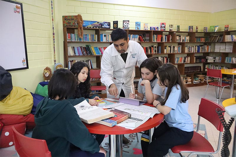 A teacher in a white lab coat guides a group of students working at a round table in a school library filled with bookshelves.