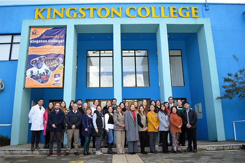 A group of staff and faculty pose for a photo in front of the bright blue entrance to Kingston College, beneath a large welcome banner.