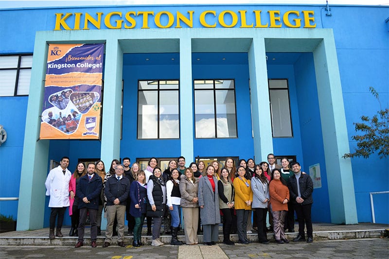 A group of staff and faculty pose for a photo in front of the bright blue entrance to Kingston College, beneath a large welcome banner.