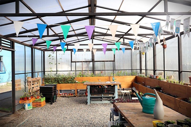 A festive greenhouse with raised garden beds, colorful triangular flags hanging from the ceiling, and various gardening tools on wooden tables.