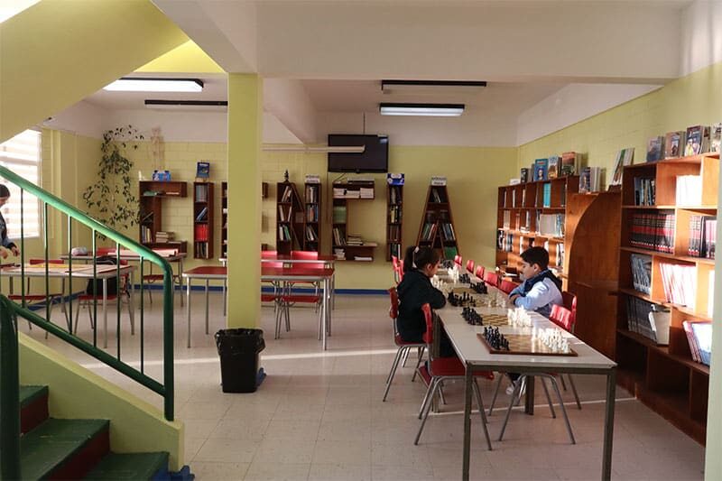 Two students play chess at a table in a school library filled with bookshelves, red chairs, and study desks.
