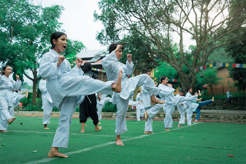Children practicing martial arts in white uniforms on a green outdoor field.