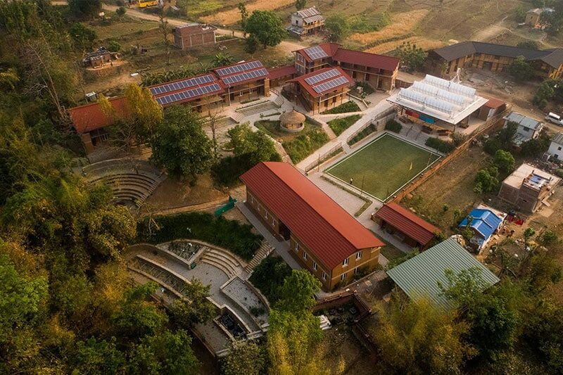 Aerial view of a rural school campus with red-roofed buildings, solar panels, an open courtyard, and outdoor amphitheaters surrounded by greenery.