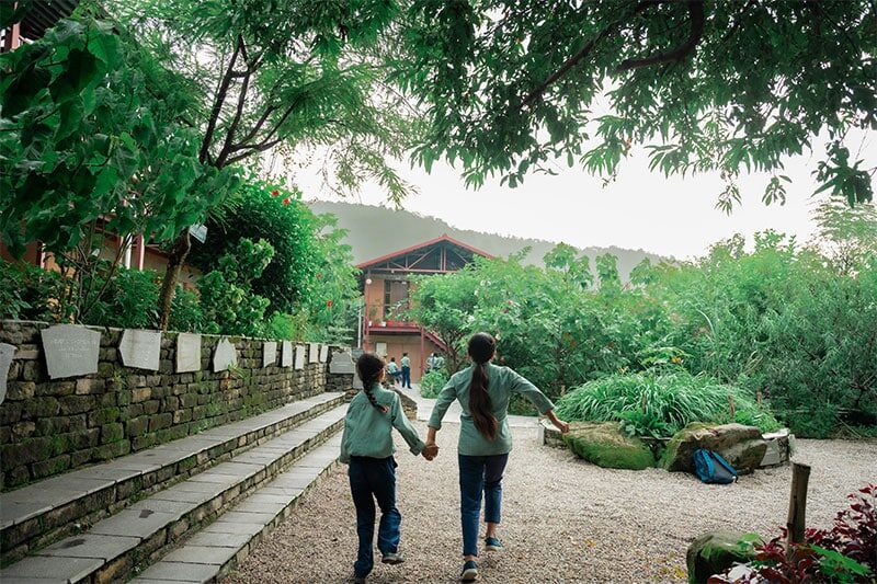 Two schoolchildren walk hand-in-hand through a lush, tree-lined campus path toward a rustic school building nestled in greenery.