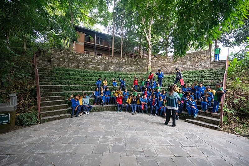 A large group of students in colorful uniforms sit on stone steps in a forested outdoor amphitheater as a teacher stands nearby.