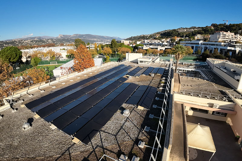 A rooftop covered with black solar panels, set against a backdrop of trees, buildings, and distant hills under a clear blue sky.