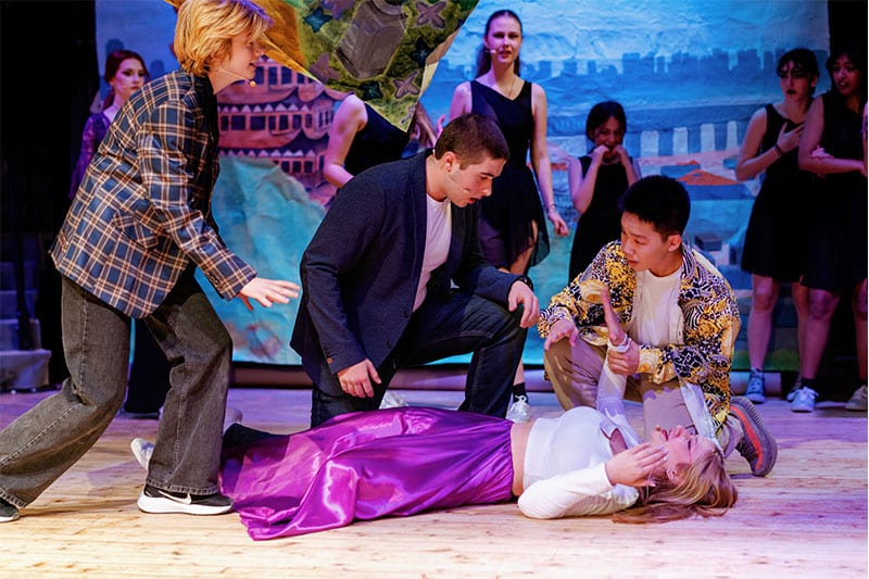 Students performing a dramatic scene on stage, with one girl lying on the floor surrounded by concerned classmates, as part of a theatrical production.