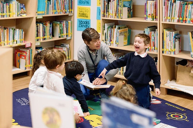 A teacher reads to a group of young children sitting on a colorful rug in a library, while one child laughs joyfully.