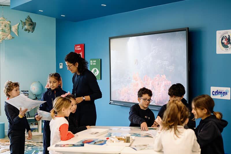 A teacher guides young students in a brightly colored classroom with a digital screen displaying underwater scenes and educational materials on the table.