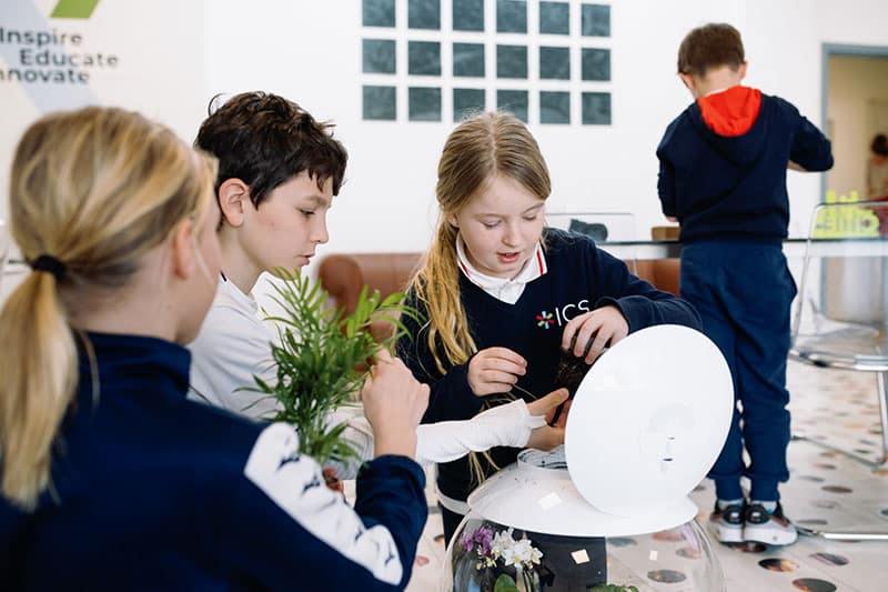 Children collaborate on a planting project using a white dome-shaped container with plants and soil, inside a modern classroom or learning space.