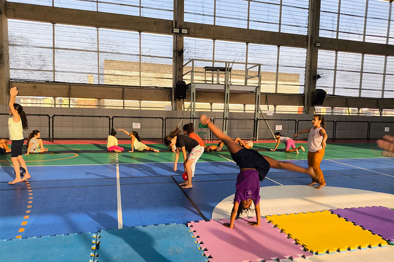 Children participating in a gymnastics or movement class in a brightly lit indoor sports hall, with one child performing a cartwheel.