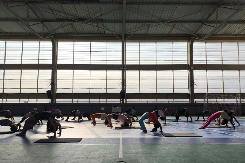 Students practising backbend yoga poses on mats inside a large indoor sports hall with natural light streaming through tall windows.