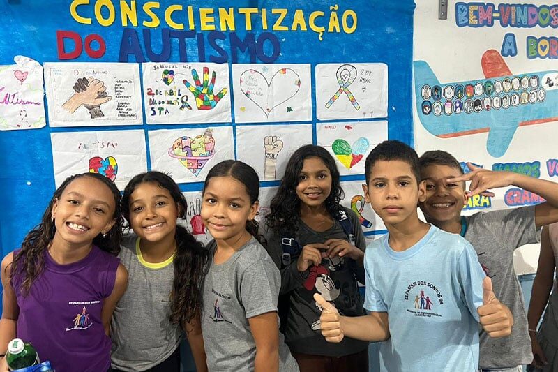 Group of smiling schoolchildren standing in front of a colourful wall display about autism awareness, featuring student artwork and messages.