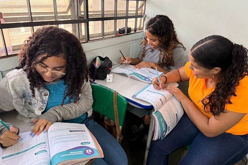 Three students sitting at classroom desks working on exercises in their textbooks, focused and writing attentively.