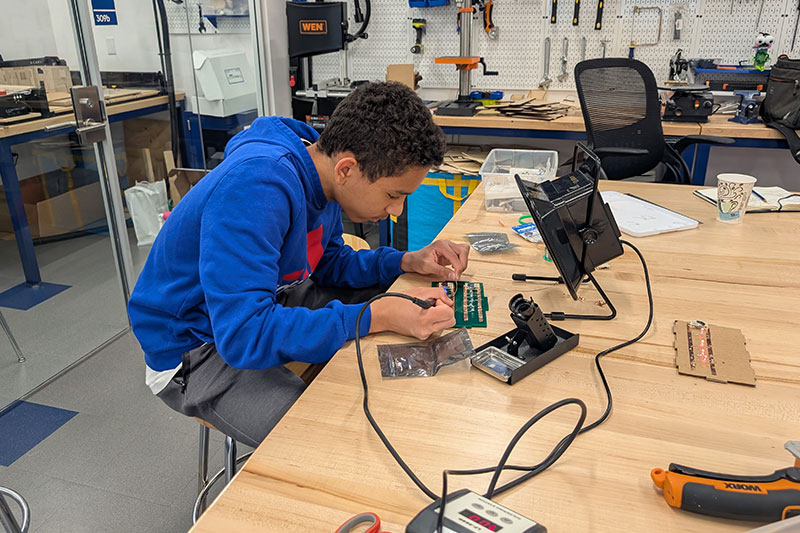 Student working on an electronics project at a workbench in a modern makerspace or tech lab.