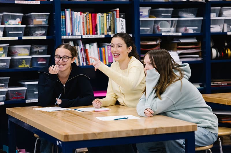 Three students sitting at a table in a classroom, smiling and pointing, with books and supply bins in the background.