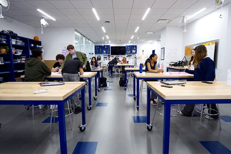 Students working in a bright, modern classroom with large workbenches, blue accents, and shelves of supplies.