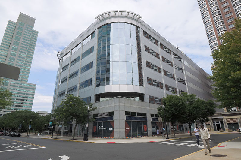 Modern multi-story building with glass and steel façade located on a city corner, surrounded by high-rise buildings.