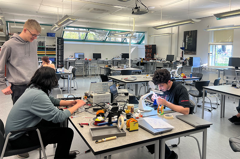 Students working with electronics and lab equipment in a spacious, well-lit technology classroom.