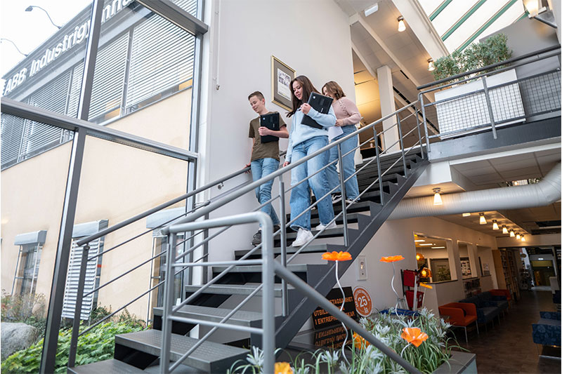 Students walking down a modern staircase inside a bright educational building, holding laptops and notebooks.