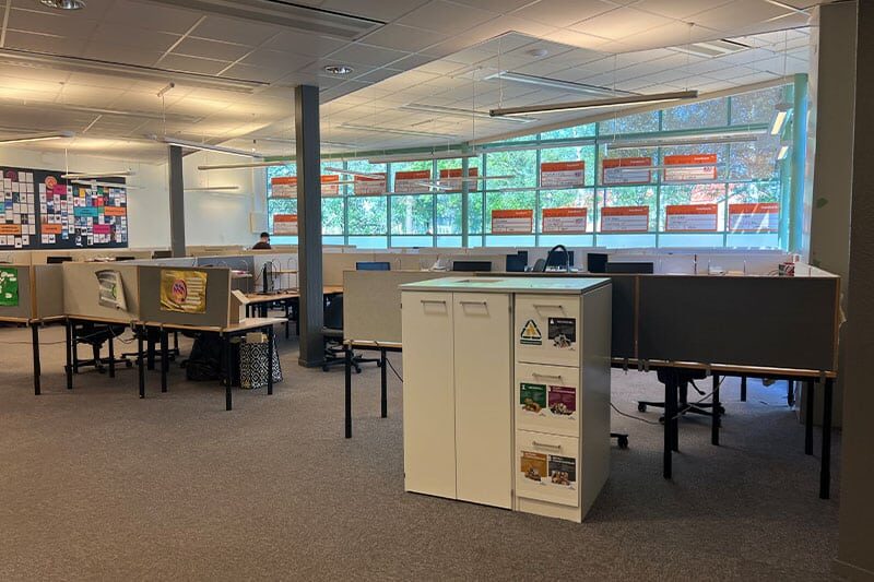 Modern open-plan study area with individual cubicles, computers, large windows, and educational posters on the walls.