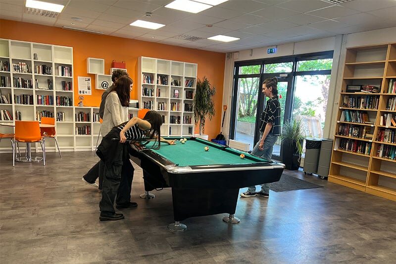 Students playing pool in a library or student lounge area with bookshelves, seating, and large windows.
