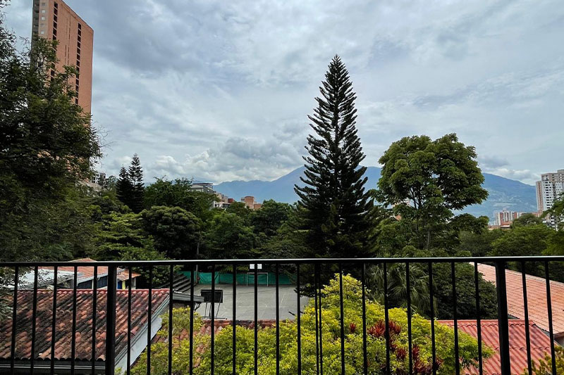 Scenic view from a balcony overlooking a tree-filled school campus with a sports court, set against a backdrop of mountains and city buildings.