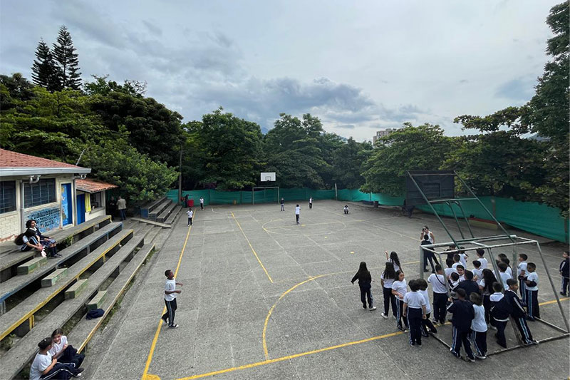 Students in school uniform gathered around a basketball court during a PE lesson or break time, with tiered seating and lush trees surrounding the area.