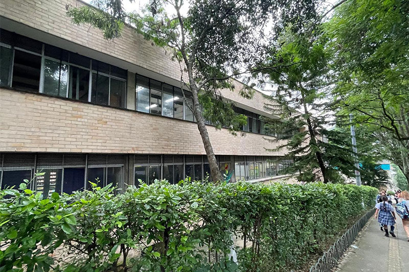 Exterior view of a modern school building with large windows, surrounded by trees and greenery, with students walking along a path.