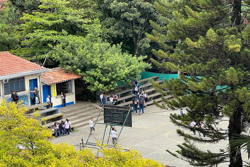 Outdoor view of a school courtyard surrounded by trees, with students gathering near a small building and seating steps, and others playing on a sports court.