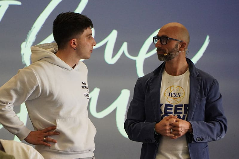 Two men engaged in a serious conversation on stage, both wearing microphones, with one in a hoodie and the other in a blazer over an IEXS Foundation t-shirt.