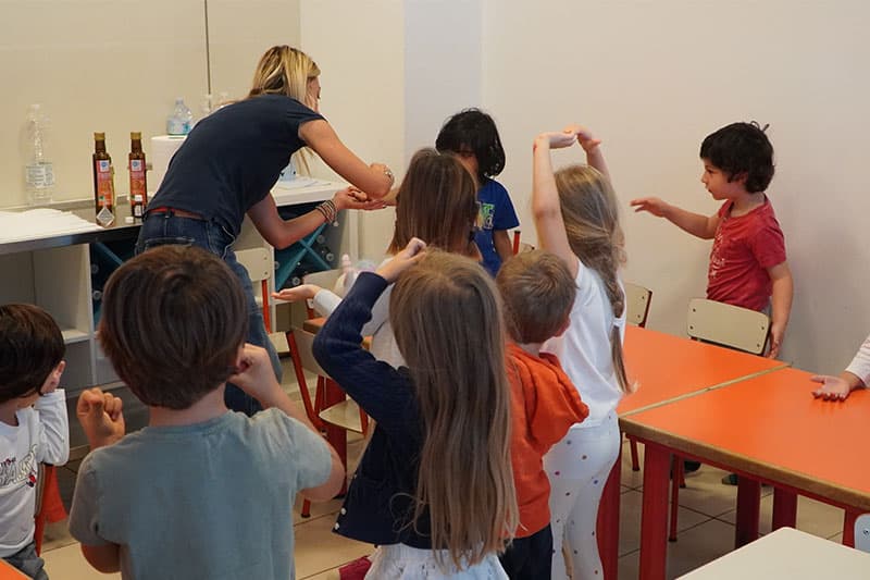 A group of young children in a classroom interact with a teacher who is guiding an activity near a counter.