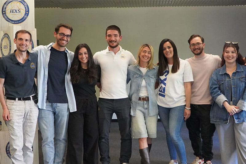 A group of eight smiling adults stand close together posing for a group photo, some wearing IEXS-branded clothing.