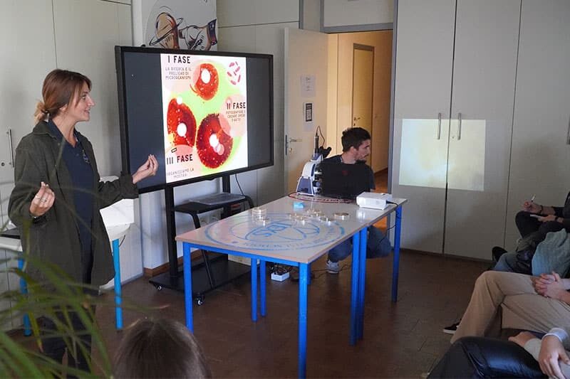 A woman presents in front of a screen showing microscopic images, while a man sits near a microscope at a table, and a small audience watches attentively.