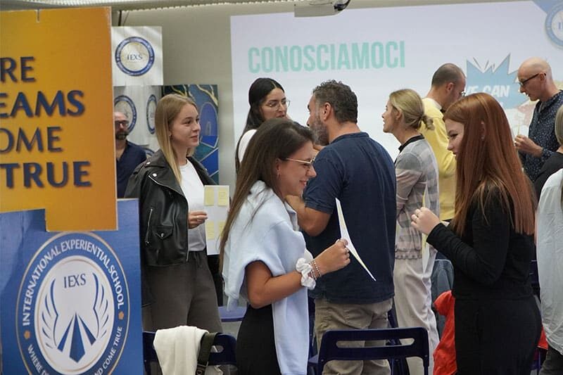 A group of adults engaged in conversation during a networking or introduction event at the International Experiential School (IEXS), with visible signage and presentation screens.