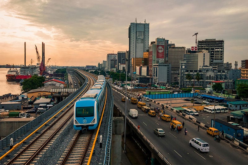 Modern blue train on an elevated railway track in a bustling city with high-rise buildings and traffic below.