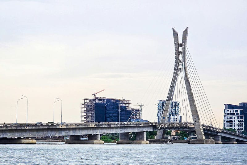 Cable-stayed bridge over water with high-rise buildings and construction cranes in the background.