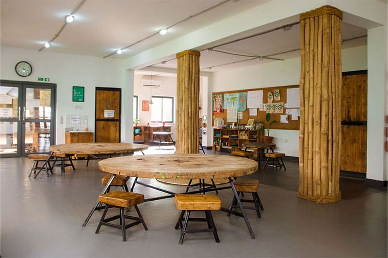 Spacious classroom or learning area with round wooden tables, stools, bamboo pillars, and educational displays on the wall.
