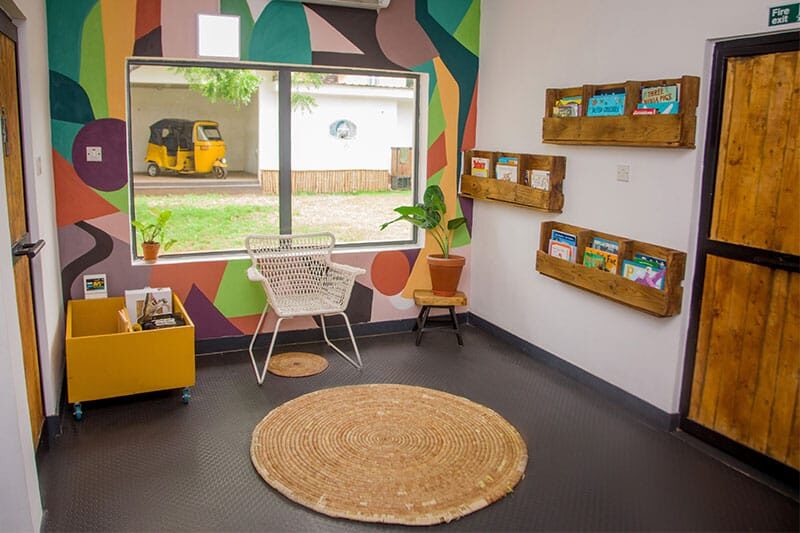 Cosy reading corner with a chair, bookshelves, potted plants, and a large window showing a yellow auto rickshaw outside.
