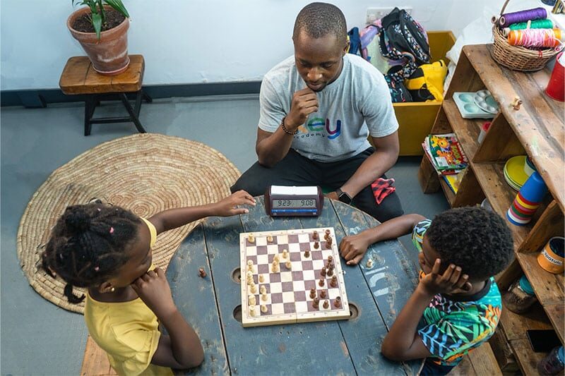 Two children playing chess at a wooden table while an adult observes thoughtfully.