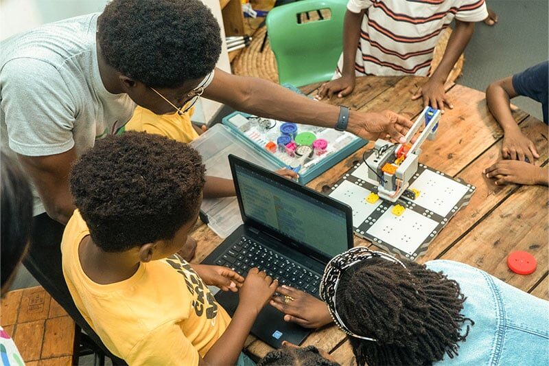 Children gathered around a laptop and robotic setup while an instructor guides them in a hands-on STEM activity.