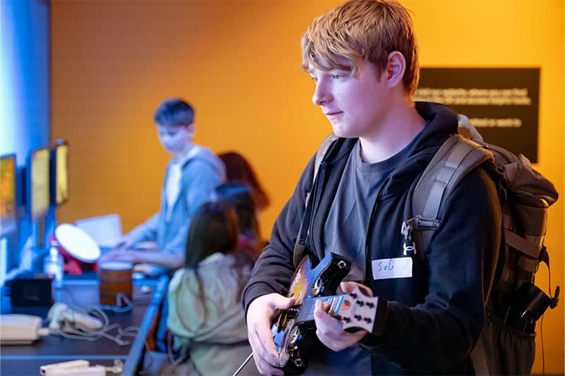 Teenager wearing a backpack playing a video game with a guitar controller, while others use computers in the background.