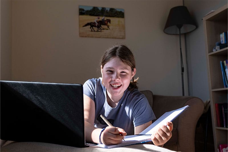 Smiling girl sitting on a couch with a laptop and notebook, engaged in online learning.