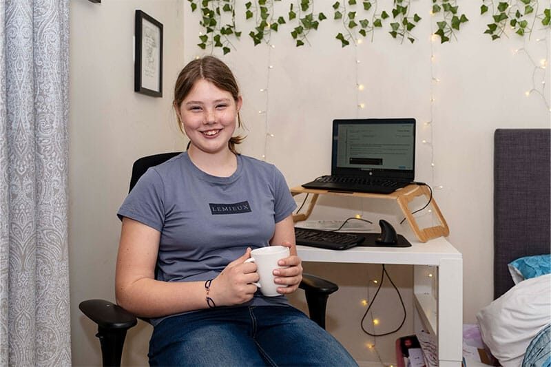 Smiling girl sitting in a home workspace holding a mug, with a laptop and keyboard on a white desk behind her.