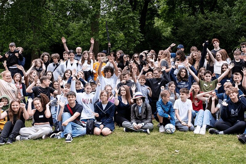 Large group of smiling students and adults posing together on a grassy field, with hands raised and trees in the background.